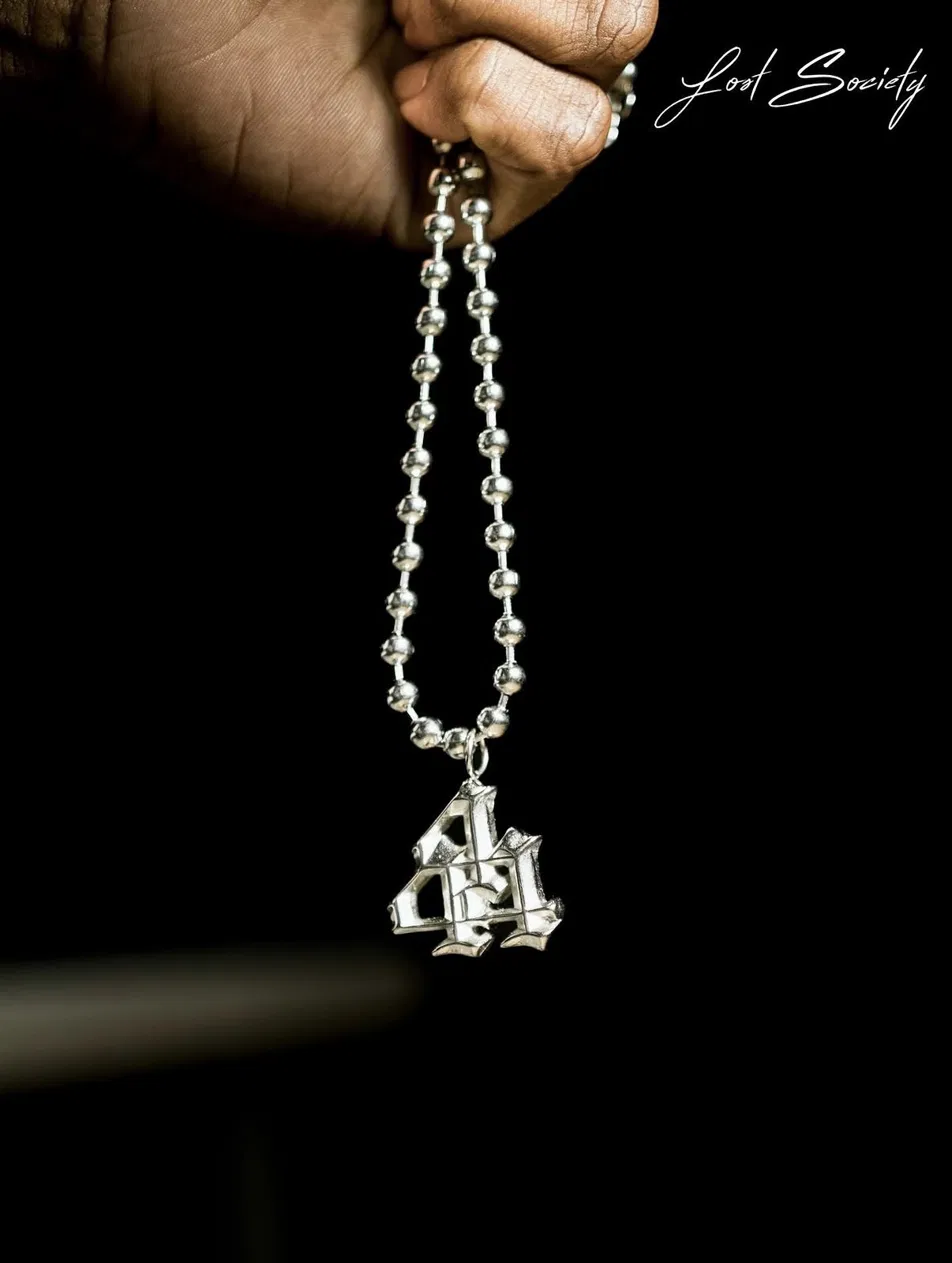 Close-up of a hand holding a silver ball-chain necklace with a blackletter 44 pendant against a black background