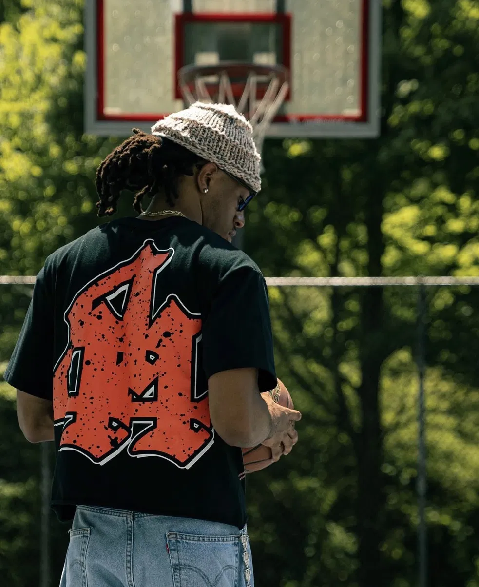 Model from behind wearing a black Lost Society tee with a large orange distressed 44 print, standing on a basketball court