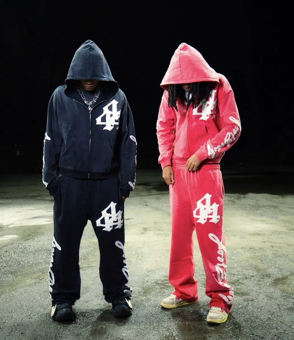 Two figures in navy and pink Lost Society tracksuits, hoods up, standing in an empty lot at night