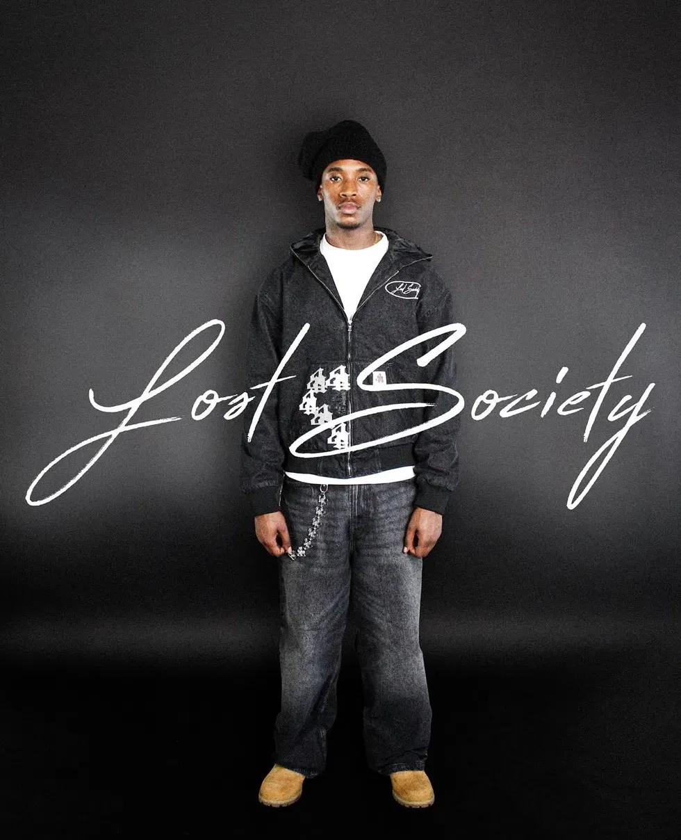 Model in a black Lost Society jacket and beanie standing against a dark backdrop with the Lost Society script logo overlaid