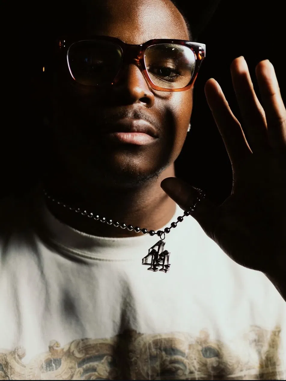 Close portrait of a model in tinted glasses with a silver 44 pendant, hand raised toward camera